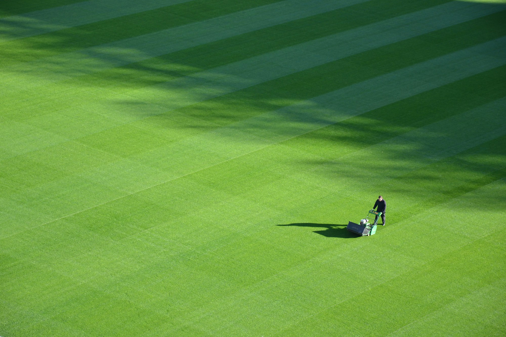 Aerial view of a large striped sports field with a single groundskeeper pushing a mower across it